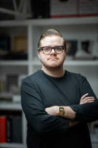 MBA team member headshot featuring a blonde male architect with short hair and beard wearing thick glasses and a long sleeve black t-shirt standing with arms crossed in front of a shelf in an office with white walls