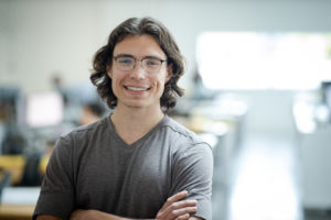 MBA team member headshot featuring a young male architect with curly brown hair, glasses, and a gray v-neck shirt standing with his arms crossed and smiling in front of a white office space